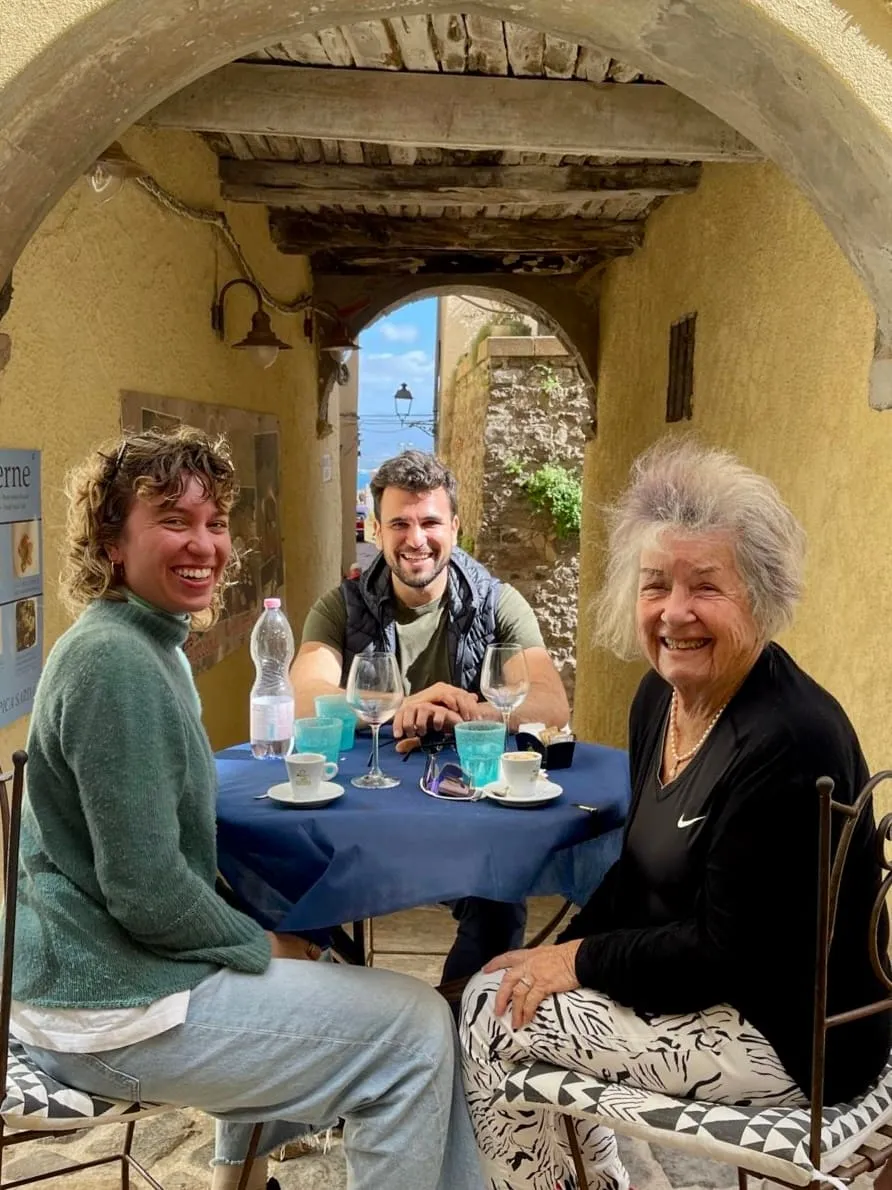 Three people are sitting at a table in a cozy alleyway under an old, rustic archway. On the left is a woman with curly hair wearing a green sweater, in the middle is a man with a beard and a vest, and on the right is an older woman with gray hair, smiling at the camera. The table has empty wine glasses, espresso cups, and a water bottle on it. The atmosphere is relaxed and friendly, with a Mediterranean vibe in the background.